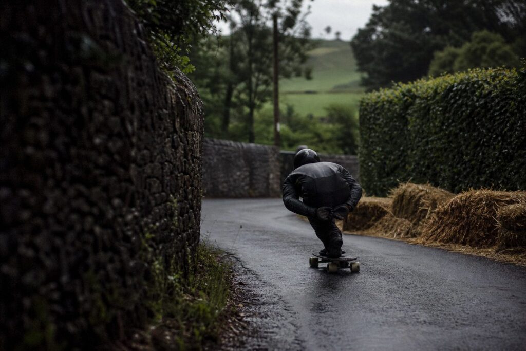 A helmeted downhill rider tucked through a narrow English country lane lined with stone walls