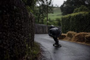 A helmeted downhill rider tucked through a narrow English country lane lined with stone walls