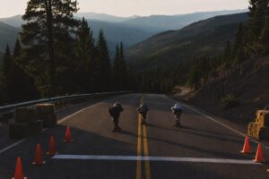 Three helmeted downhill longboarders tucked low on an empty mountain highway at dawn, hay bales and cones lining the start line